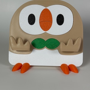 Puede incluir: Un soporte con forma de búho en color beige y blanco, con grandes parches oculares blancos, pupilas negras, pico naranja, un detalle verde en forma de pajarita y dos pies naranjas. Incluye dos soportes beige de tres puntas en el frente.