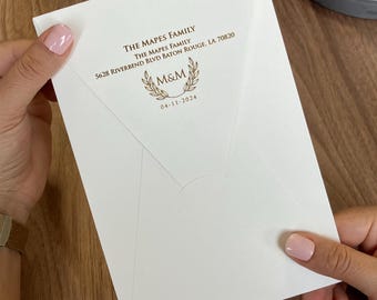 以下が含まれることがあります： 白い封筒には、金色の文字で「The Mapes Family, 5628 Riverbend Blvd Baton Rouge, LA 70820」と印刷されており、2本の月桂樹の枝に囲まれたM&Mのモノグラムと、その下に日付「04-11-2024」が記されています。