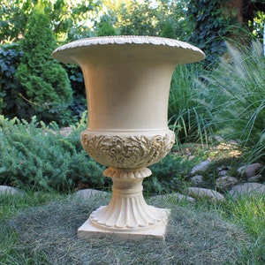 Puede incluir: Un macetero grande, de color crema, adornado con una base ancha y un diseño floral decorativo. El macetero está sentado en un lecho de hierba verde.