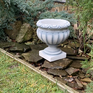 May include: A grey concrete planter with a fluted design and a square base. The planter is sitting on a bed of rocks.