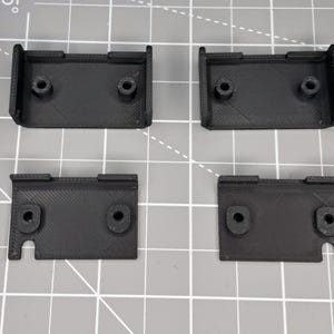 May include: Four black plastic brackets with holes for screws. The brackets are designed to be mounted to a surface.