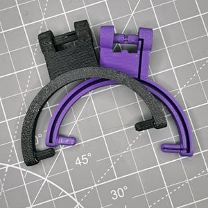 May include: Two 3D-printed objects. One is black, the other is purple. Each has a rectangular top and a curved, semi-circular base. The black object has a textured surface. The purple object has a smooth surface. The objects are on a gray surface.