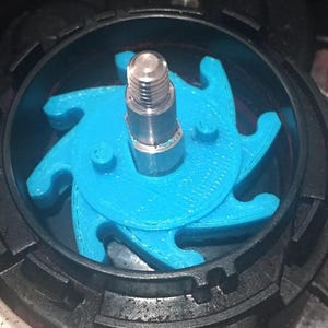 Puede incluir: Primer plano de un componente mecánico. Un impulsor azul brillante, con múltiples lóbulos, se encuentra dentro de una carcasa negra. Un perno de metal plateado está centrado. El diseño sugiere una bomba o un mecanismo similar.