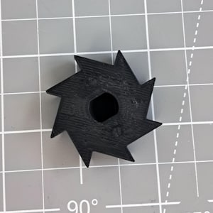 Könnte beinhalten: Ein schwarzes, sternförmiges Objekt mit scharfen, spitzen Kanten und einem sechseckigen Loch in der Mitte. Das Objekt scheint aus einem festen Material zu bestehen und befindet sich vor einem grauen Rasterhintergrund.
