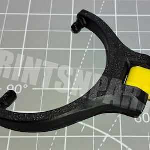 May include: A black, Y-shaped mechanical component with a textured surface, featuring two curved arms and a central hinge. A bright yellow, cylindrical plastic part is attached to the hinge, extending outward from the black frame.