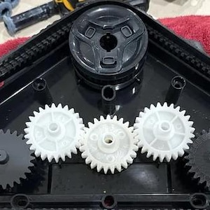 Può includere: Primo piano di un componente robotico in plastica nera con più ingranaggi e ruote. Gli ingranaggi sono bianchi e neri e le ruote sono nere. Il componente è su una superficie rossa. Parti meccaniche.