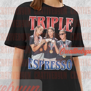 Könnte beinhalten: Schwarzes Unisex-T-Shirt mit den Worten "TRIPLE ESPRESSO" in roter und blauer Schrift. Das Shirt zeigt eine Grafik von drei Personen, die Espressotassen halten und Medaillen tragen. Das Shirt ist unisex.