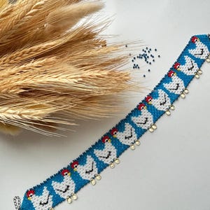 May include: A beaded bracelet with a blue background and white chicken pattern. The chickens have red combs and yellow beaks. The bracelet has a silver clasp and small gold-colored dangles.