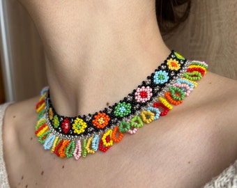 Może przedstawiać: Kolorowy naszyjnik choker z koralików z czarną podstawą i frędzlami z wielokolorowych koralików. Naszyjnik ma wzór małych, kolorowych kwiatów.