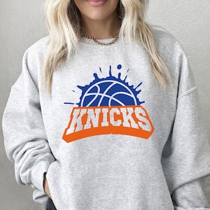 May include: A light gray sweatshirt featuring the New York Knicks logo. The logo includes a blue basketball design with orange text that reads "KNICKS". The sweatshirt is a crew neck style.