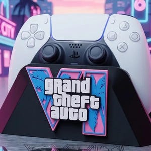 Op de afbeelding: Een wit en zwart videospelcontroller in een zwarte standaard. De standaard bevat een roze en blauwe afbeelding met de tekst Grand Theft Auto en een groot Romeins cijfer VI, tegen een gestileerde neonverlichte stedelijke achtergrond.
