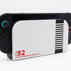 Peut inclure: Une console Nintendo Switch noire insérée dans une station de charge blanche. La station de charge présente un design rappelant la console Nintendo Entertainment System classique, avec le texte "Nintendo 2 Entertainment System" en rouge.