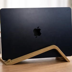 May include: A dark blue laptop with a centered Apple logo is positioned upright in a light-colored wooden stand. The stand features an angled, minimalist frame that supports the device in a vertical orientation, providing a stable base.