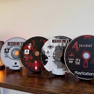 May include: Four Resident Evil PlayStation game discs on a wooden shelf. The discs have game titles and graphics in black, red, and white. Each disc is held by a small, dark brown stand.