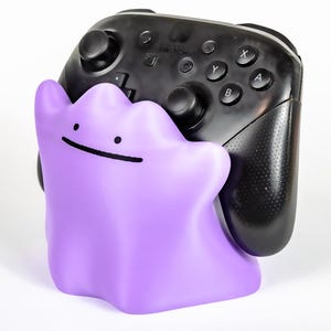 Puede incluir: Un soporte para mando de Nintendo Switch con forma de Ditto morado. El soporte sujeta un mando Switch Pro negro. El mando tiene los botones A, B, X e Y. La figura de Ditto tiene una simple sonrisa negra.
