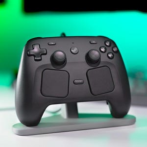 May include: A matte black video game controller featuring two analogue sticks, a directional pad, four action buttons, two large touchpads, and a central Steam logo button, resting on a light grey plastic stand with a horizontal base.