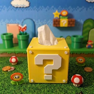 May include: A yellow cube-shaped tissue box cover featuring a white question mark on the front face. The box has small white circular accents on the corners and a single white tissue protruding from the top opening, designed to resemble a game block.