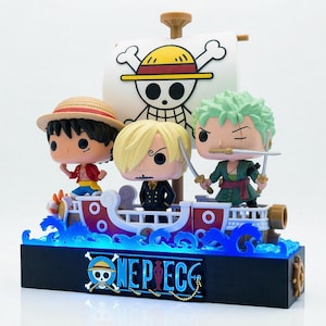 Può includere: Un oggetto da collezione di One Piece con tre personaggi su una nave con una vela a teschio e ossa incrociate. La base ha il logo di One Piece ed emette luce blu. Le figure includono Luffy, Sanji e Zoro.