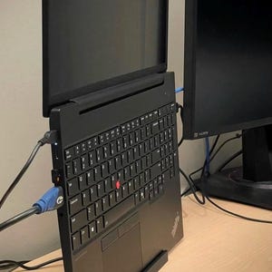May include: A black ThinkPad laptop positioned vertically in a black stand, featuring a QWERTY keyboard with a red pointing stick, a trackpad, and various ports connected to cables, placed next to a secondary black computer monitor.