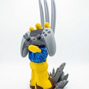 May include: A yellow and blue gloved hand sculpture featuring three long silver claws extending upward. The hand holds a gray wireless video game controller. At the base of the sculpture, there is a cluster of jagged gray rock-like formations.