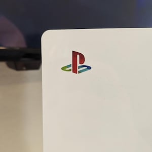 May include: A white gaming console side panel featuring a multi-coloured PlayStation logo in the upper corner. The logo displays a gradient of red, yellow, green, and blue colours, set against the smooth, matte white surface of the hardware casing.