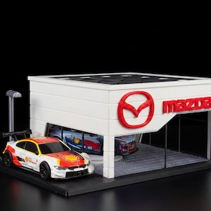 Könnte beinhalten: Ein weißes Mazda-Autohaus-Modell mit rotem Mazda-Logo und Schriftzug an der Fassade. Ein weiß-rot-gelber Rennwagen im Shell-Design steht davor, während ein weiterer blau-roter Rennwagen im Inneren des Gebäudes zu sehen ist.