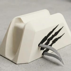 May include: A cream-colored, wedge-shaped desk organizer or stand featuring a decorative black claw mark graphic on the side. The object has a central recessed slot and two small, grey, curved plastic prongs extending from the lower right side.