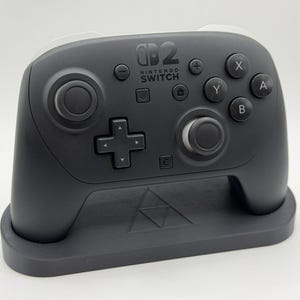 May include: A matte dark gray wireless game controller for Nintendo Switch resting on a matching dark gray stand. The controller features a central D-pad, two analog sticks, four action buttons labeled A, B, X, Y, and the Nintendo Switch logo.