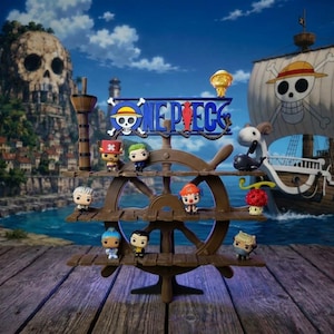 Puede incluir: Un expositor marrón con forma de timón de barco que cuenta con tres estantes horizontales con varias figuras pequeñas de vinilo. En la parte superior del soporte aparece el logotipo azul de One Piece. El soporte se sitúa sobre un muelle de madera.