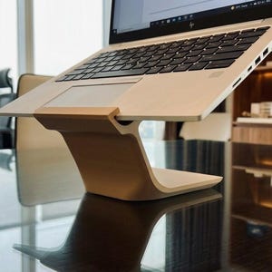 Può includere: Un computer portatile è posizionato su un supporto per laptop in legno chiaro. Il supporto ha un design curvo ed è posizionato su una superficie nera riflettente. Il laptop è aperto, mostrando uno schermo, e lo sfondo include mobili per ufficio e una finestra.