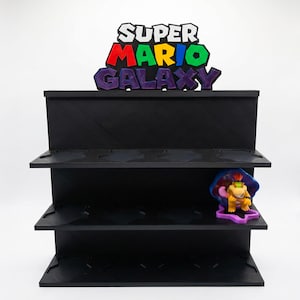 Puede incluir: Estantería negra de varios niveles con las palabras "SUPER MARIO GALAXY" en letras coloridas. Una pequeña figura de Bowser Jr. está en el estante inferior. La estantería está diseñada para guardar objetos de colección.