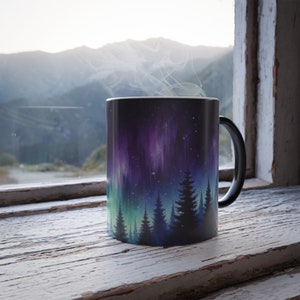 May include: A black coffee mug with a colorful aurora borealis design and a silhouette of a forest. The mug is sitting on a white wooden surface with a window and mountains in the background.
