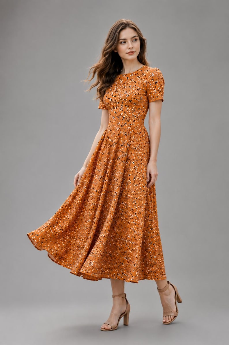 May include: A midi-length dress in a burnt orange hue featuring a dense, speckled pattern of small white and black dots. The garment has a round neckline, short sleeves, and a fitted bodice that transitions into a full, flared skirt.