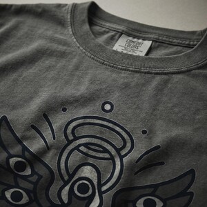 May include: Dark gray Comfort Colors t-shirt featuring a graphic design of an angel with multiple eyes and an halo. The shirt has a crew neck and a tag that reads "Comfort Colors". The graphic is in dark blue.