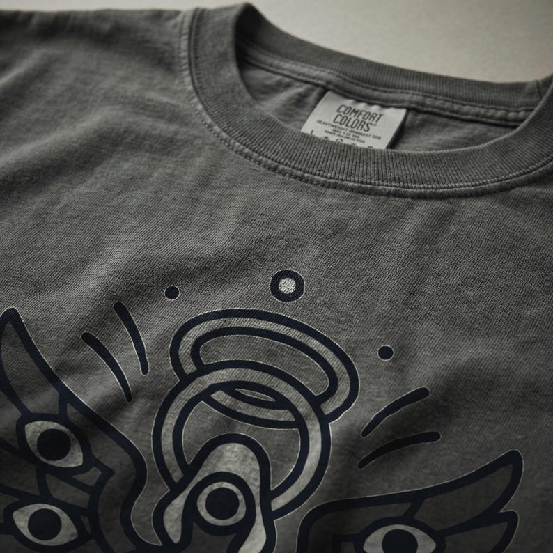 May include: Dark gray Comfort Colors t-shirt featuring a graphic design of an angel with multiple eyes and an halo. The shirt has a crew neck and a tag that reads "Comfort Colors". The graphic is in dark blue.