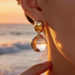 Puede incluir: Pendientes dorados con una gota esférica transparente. Los pendientes tienen una parte superior redondeada y una parte inferior redonda transparente. El sol poniente se refleja en el orbe transparente. Un accesorio elegante.