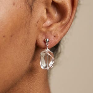 May include: A silver stud earring featuring a dangling, clear, irregular-shaped gemstone. The stone is suspended from a small silver ball stud by a delicate wire wrap, showcasing a transparent, faceted appearance that catches the light.