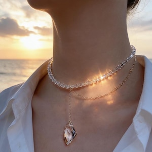Puede incluir: Un collar de varias capas con un choker de cuentas de cristal transparente y una fina cadena dorada. El colgante es un cristal facetado con un engaste dorado. El collar se lleva sobre una camisa blanca.