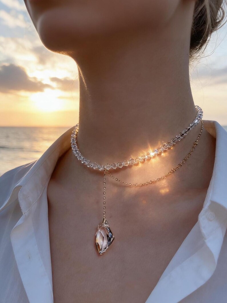 Puede incluir: Un collar de varias capas con un choker de cuentas de cristal transparente y una fina cadena dorada. El colgante es un cristal facetado con un engaste dorado. El collar se lleva sobre una camisa blanca.