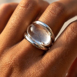 May include: A silver ring featuring a large, smooth, clear, oval-shaped cabochon stone set within a polished metallic band. The design consists of a thick, rounded silver frame that cradles the transparent, dome-shaped centre piece securely.