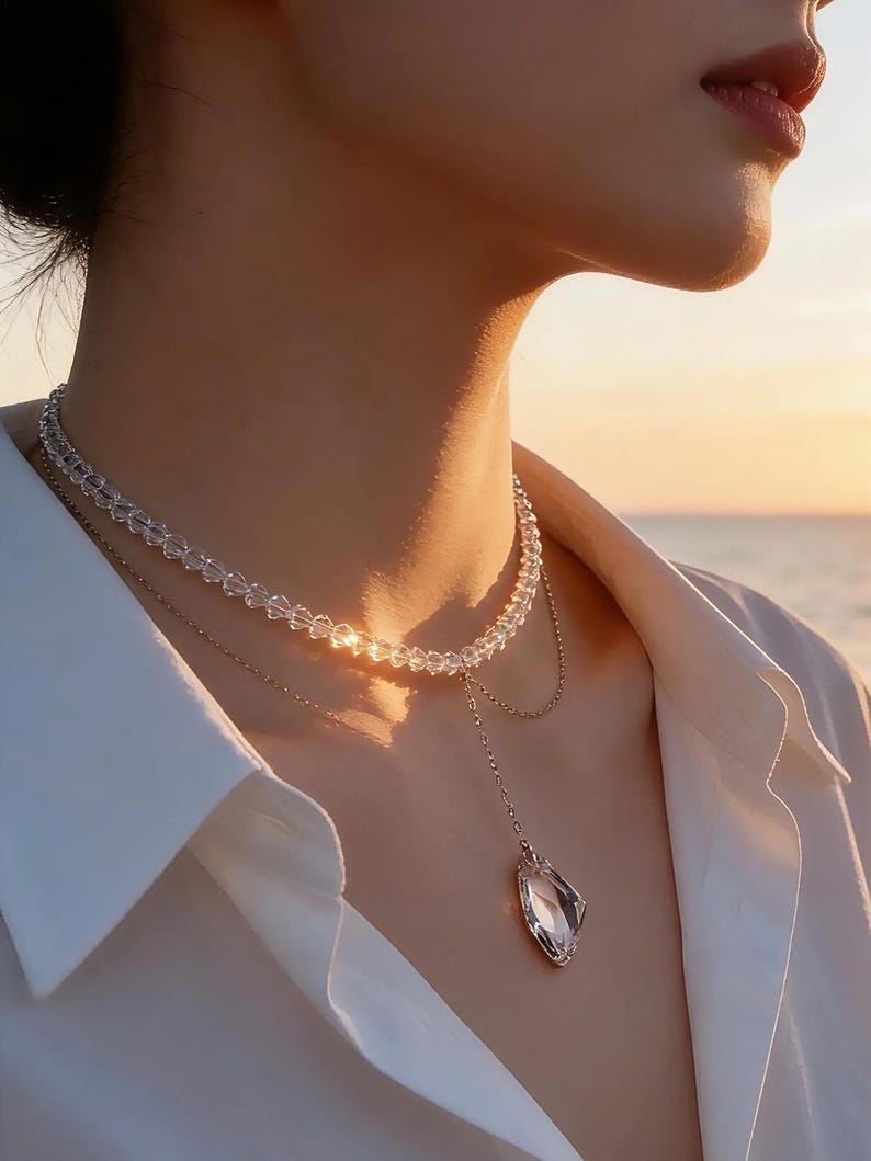 Puede incluir: Un delicado collar con cuentas de cristal transparente y un colgante. El collar tiene dos hilos, el superior compuesto por peque&ntilde;as cuentas redondas. El hilo inferior presenta un colgante en forma de l&aacute;grima. La joya se lleva sobre una camisa blanca.