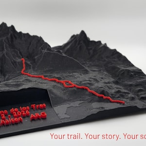 Puede incluir: Una escultura topográfica impresa en 3D de color negro de una cadena montañosa con un sendero rojo en relieve. El texto "Laguna de los Tres 22-2-2026 El Chalten ARG" está grabado en rojo en la esquina frontal. La base es un cuadrado negro plano.