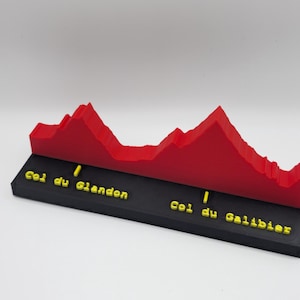 Könnte beinhalten: Ein 3D-gedrucktes Modell eines Gebirgshöhenprofils in Rot, montiert auf einem schwarzen rechteckigen Sockel. Der Sockel zeigt gelben erhabenen Text mit der Aufschrift Col du Glandon und Col du Galibier sowie kleine gelbe vertikale Markierungen.