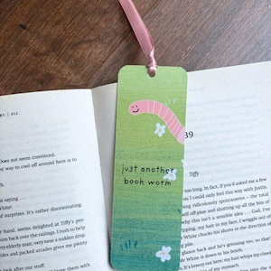 May include: A green bookmark with a pink ribbon. The bookmark features a cartoon worm with a smile and the text "just another book worm".