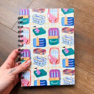 May include: A spiral-bound notebook with a repeating pattern of sardine tins in pink, blue, green, and red. Each tin is labelled "Sardines" and set against a yellow and white checkered background. The notebook is held by a hand.
