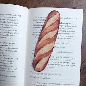 May include: A novelty bookmark in the shape of a baguette, featuring a detailed illustration of bread. The bookmark is positioned in an open book, adding a playful element to the reading experience. The book is open to page 11.