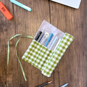May include: A green and white gingham fabric pencil roll with a pale pink lining. The roll is open and contains several pens, pencils, and a ruler.