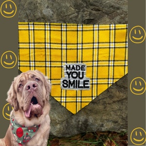 May include: A yellow plaid dog bandana with the text "MADE YOU SMILE" on a patch. The bandana is displayed with a large dog wearing a holiday-themed bandana. The background is a rock and a brown surface with yellow smiley faces.