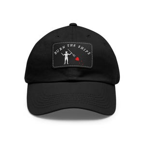 May include: Black baseball cap with a curved peak. The cap has a rectangular patch with the words "BURN THE SHIPS" and a skeleton figure holding a trident. Red heart accents are also present on the patch.