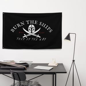 May include: Black flag with white text that reads "Burn the Ships" with a Mandalorian helmet and crossed swords underneath. The text "This is the Way" is below the helmet and swords.